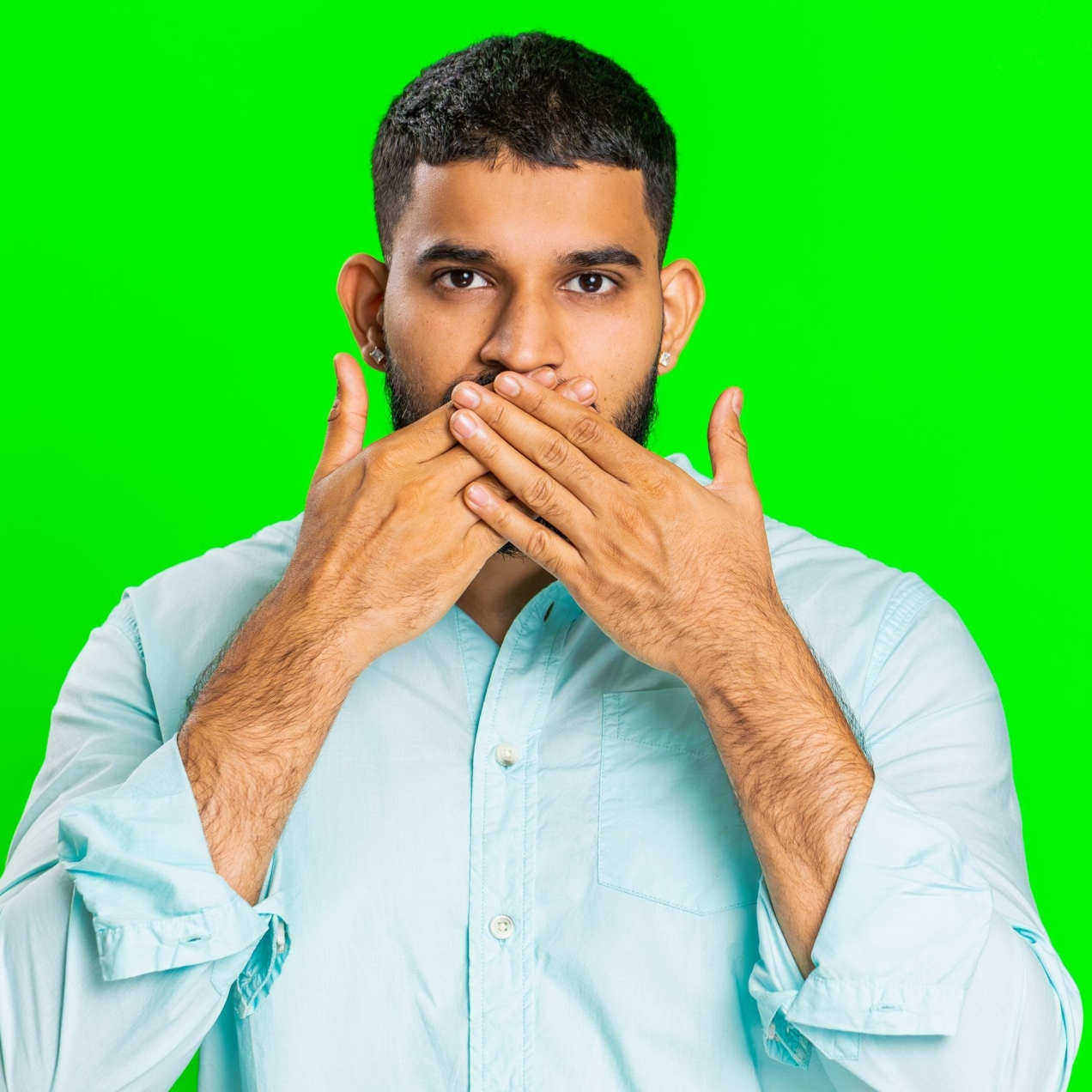 An Asian man with short dark hair hold his hands up to his mouth as if silencing himself. He wears a light blue shirt. The background is green
